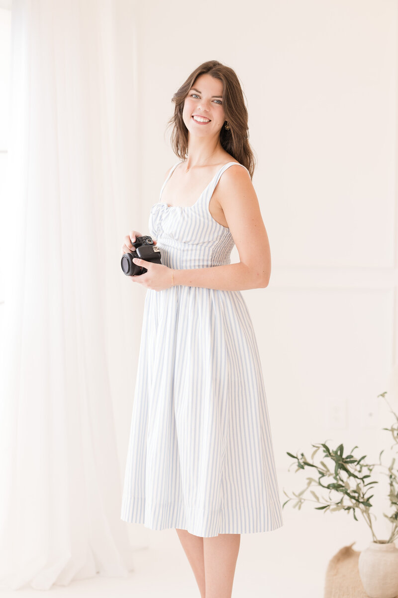 Erin standing in a bright, window-lit room in a dress and holding a camera