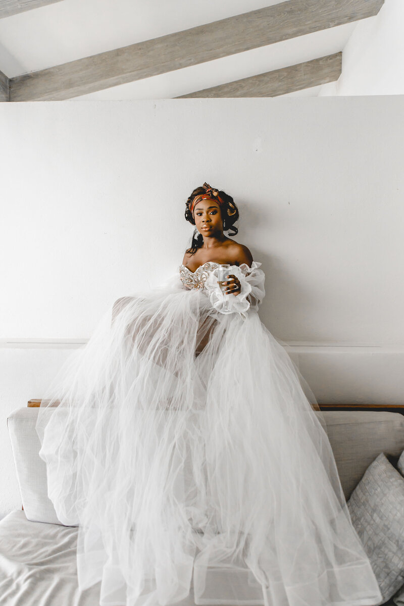 A bride in an off-the-shoulder wedding gown with voluminous tulle and lace sleeves sits on a couch against a white wall. She wears a floral crown and looks ahead, with her long gown draped elegantly around her.