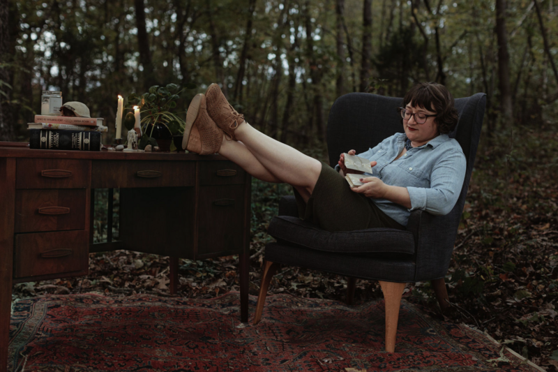 Ash sits in a chair in the forest with her feet up on a side table as she shuffles the cards of the Energy Archaeology Oracle Deck