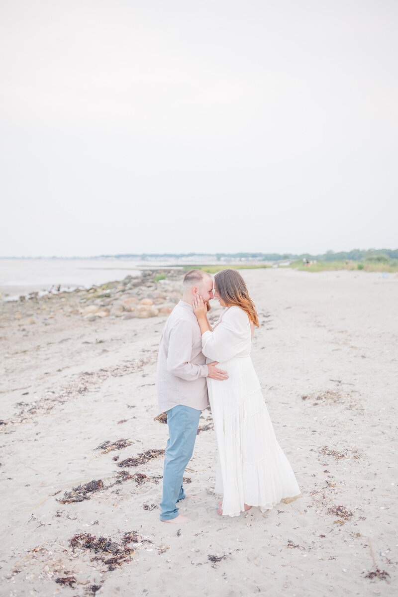 A husband and wife share a loving gaze as they stand together on the beach during a maternity session. The husband gently places his hands on his wife's belly, both smiling in the serene moment. The ocean and soft, golden light create a peaceful, romantic backdrop.