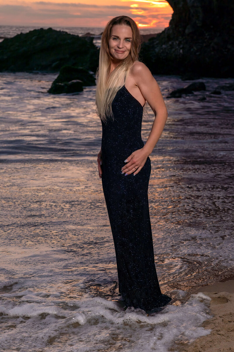 Blonde woman in elegant navy sequin dress standing on a beach at sunset, with gentle waves lapping at her feet, embodying serene beachside elegance in Laguna