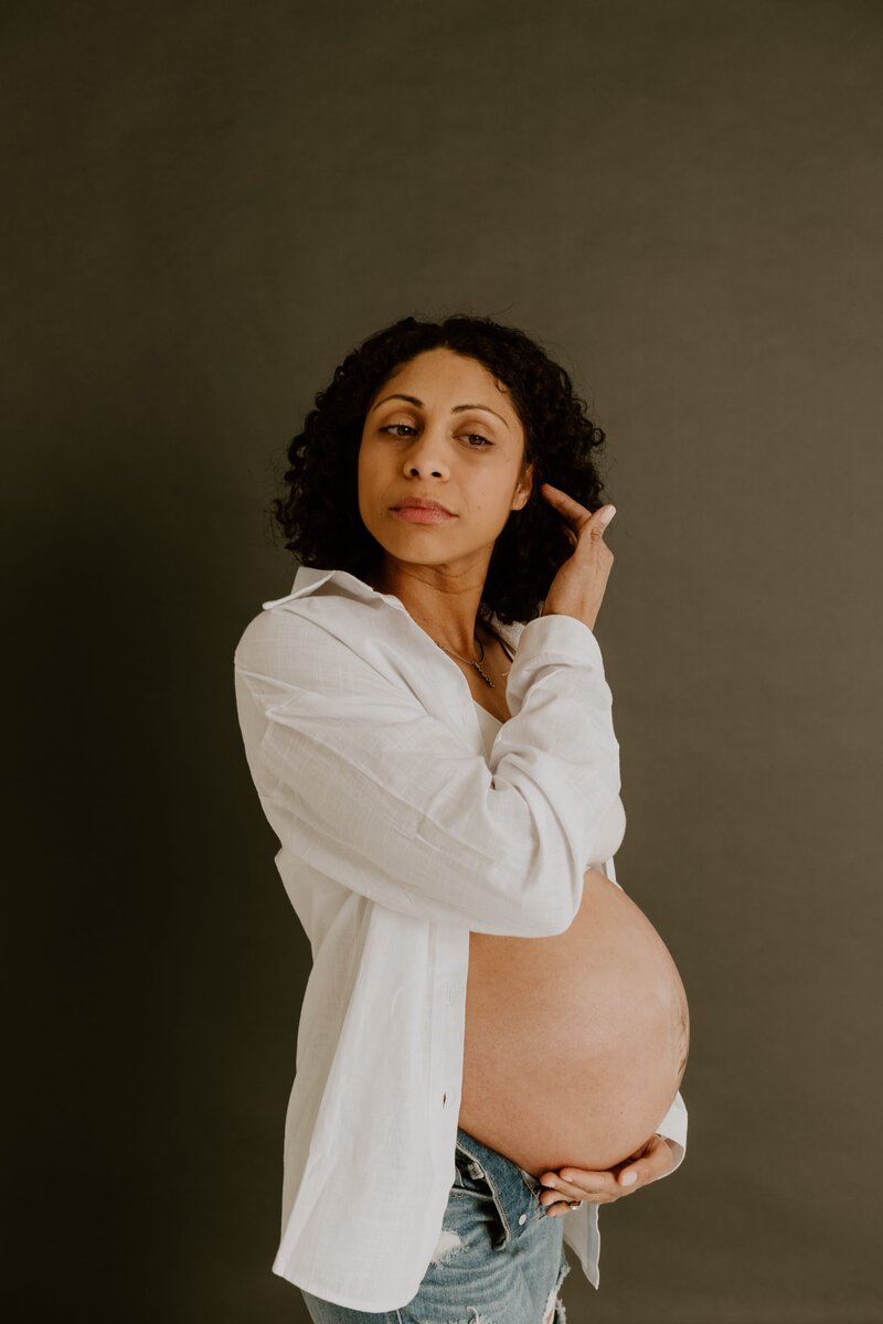 A confident pregnant woman in a white shirt and denim jeans, cradling her belly against a dark gray backdrop.