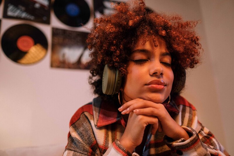 This image shows a young feminine-presenting person of color, as they listen to music on their headphones with eyes closed, head resting in their hands.
