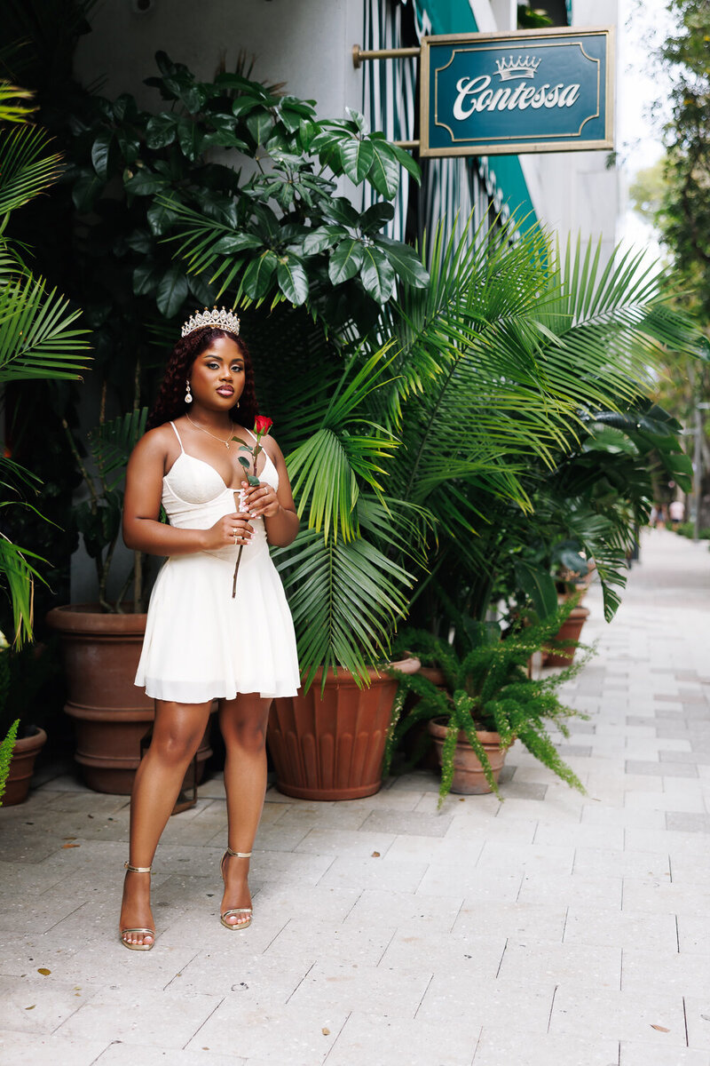 A woman in a dress holding a rose standing and posing for the camera.