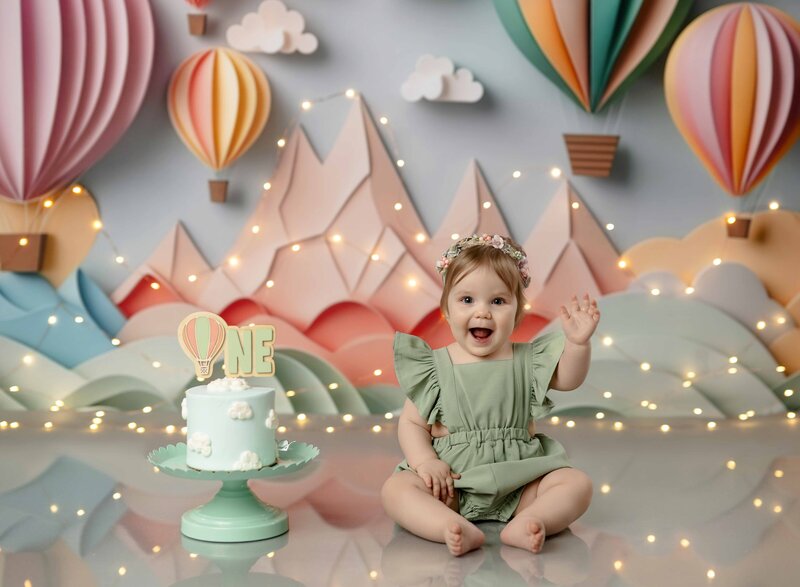 Baby girl in sage romper, waving at camera with a big smile as she prepares to get messy with her birthday cake.