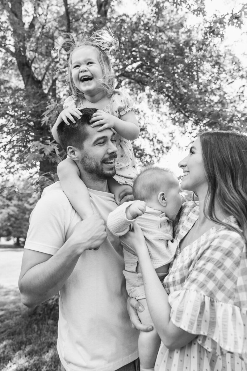 Black and white portrait of a family of four looking at one another smiling with daughter on dad's shoulders and mom holding little brother in Farmington, Missouri. 