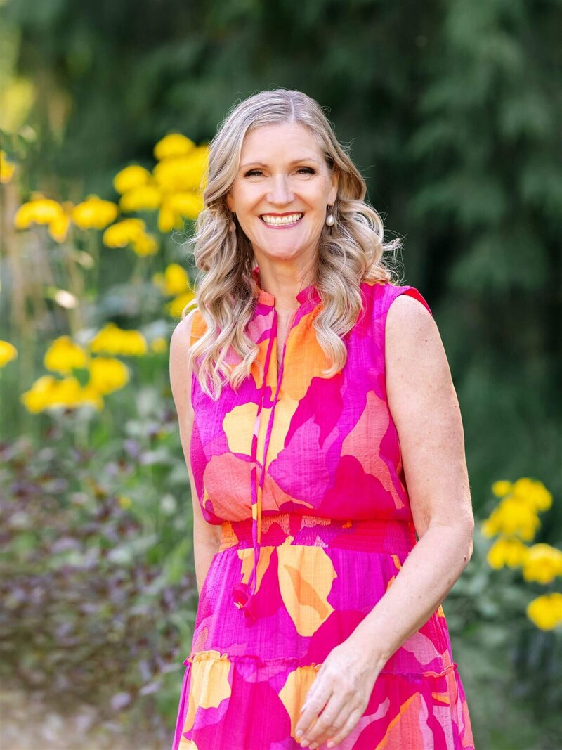 Phillipa Leseberg, an author with long blonde hair, stands outdoors in a bright pink and orange dress, surrounded by vibrant yellow flowers.