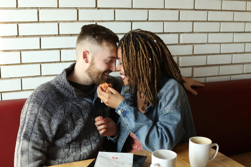 Interracial couple laughing and sharing donuts during coffee shop engagement shoot