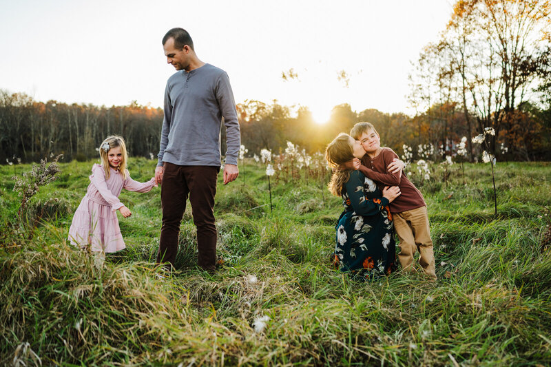 Family in a field with mom squatting and kissing son while dad is dancing with his daugther