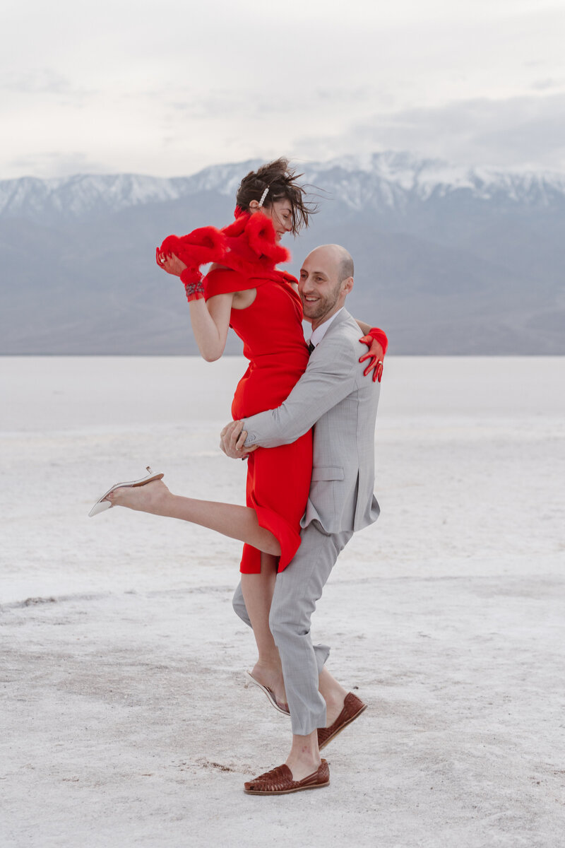 A man lifts a woman wearing a bright red dress in celebration. They are standing on white salt flats in Death Valley National Park with mountains behind them