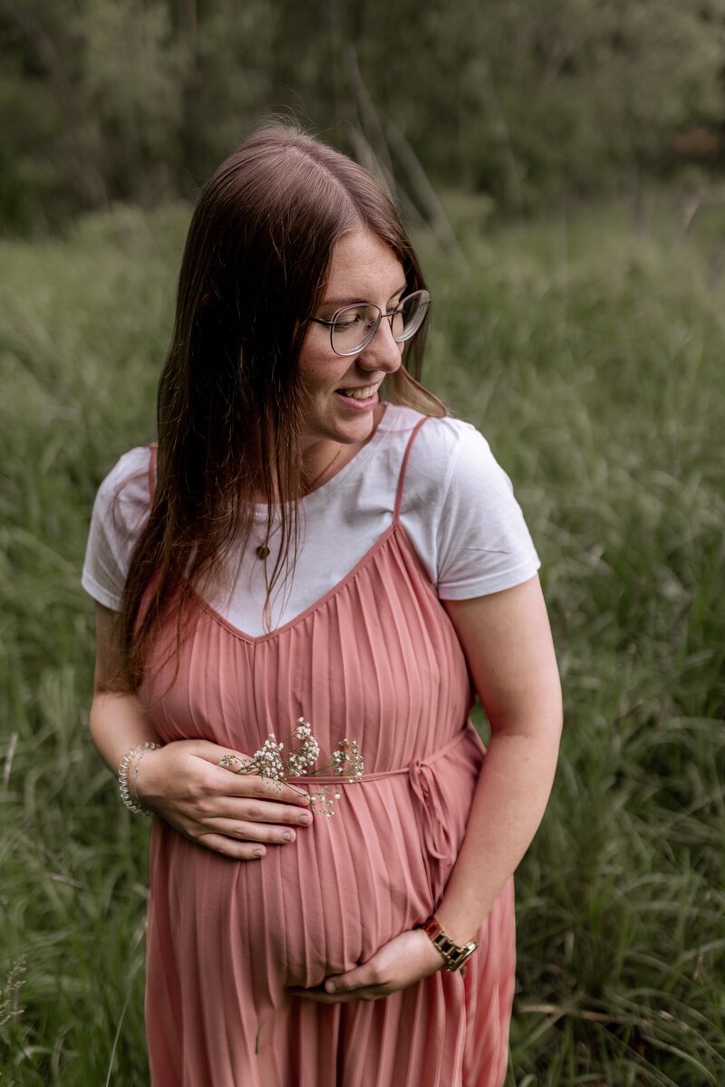 Frau in Rosa Kleid haelt Babybauch auf einer Wiese