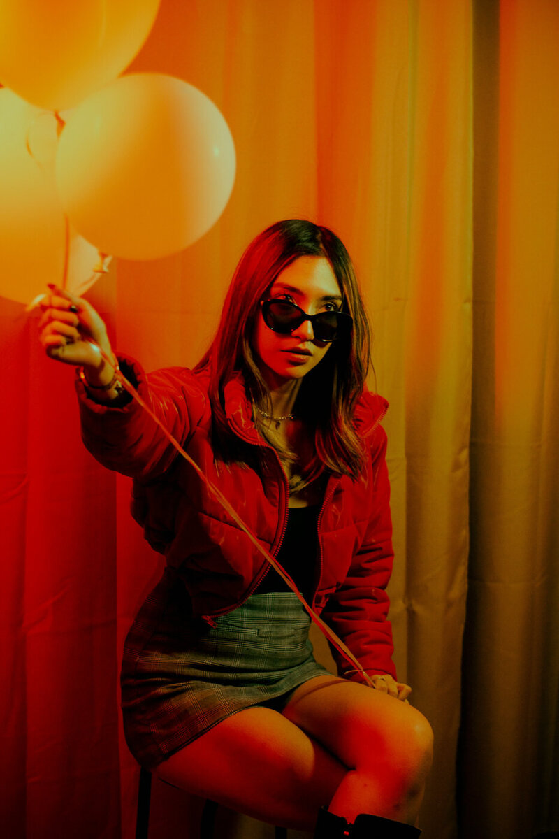 Bold woman in an orange lit room with sunglasses and a dress, holding yellow balloons.