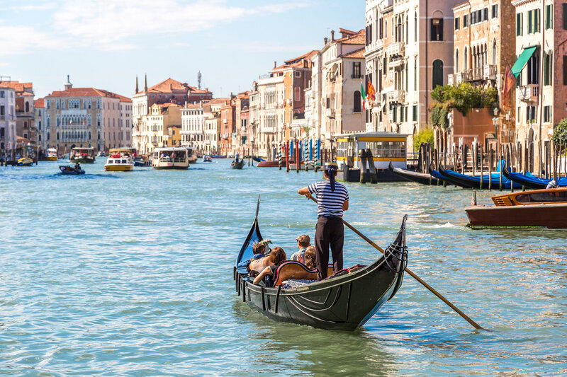 A gondolier rows a gondola with passengers along a busy canal in Venice. Historic buildings line the waterway, and several other boats are visible in the background under a clear blue sky.