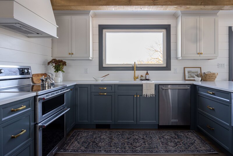 Blue cabinet kitchen centered by a window and expose ceiling beams. There is a double range on the left and peninsula on the right with white countertops and white walls.