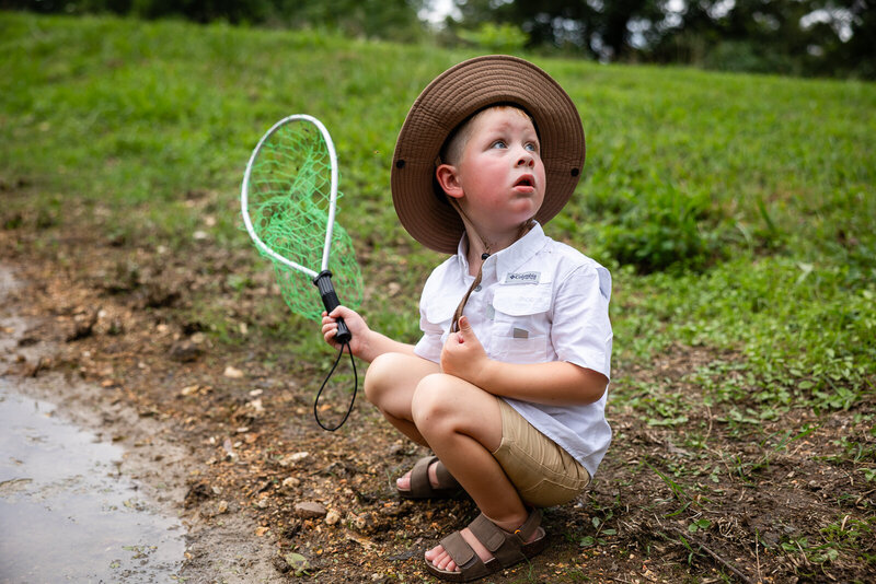 Real life documentary portrait of a little boy holding his fishing net and wearing his fishing hat by the pond. 