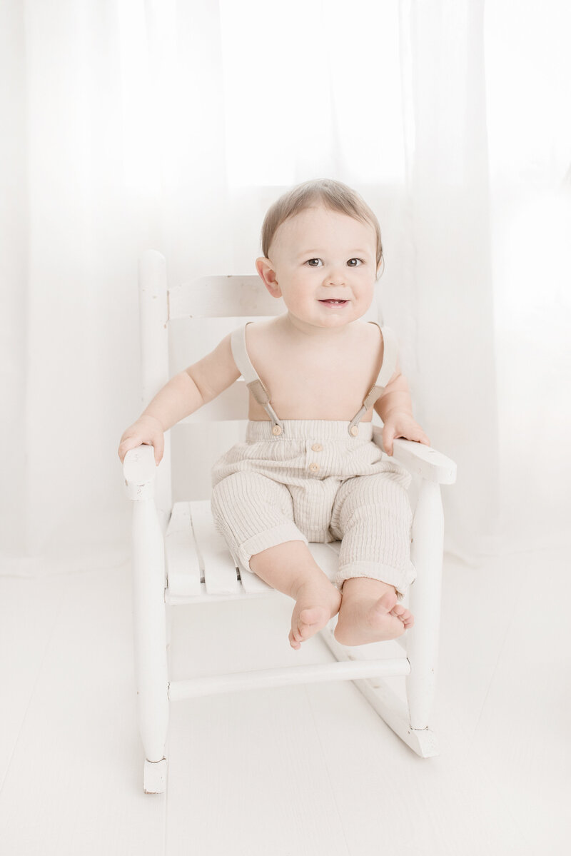 One year old boy in suspenders sitting on white rocking chair