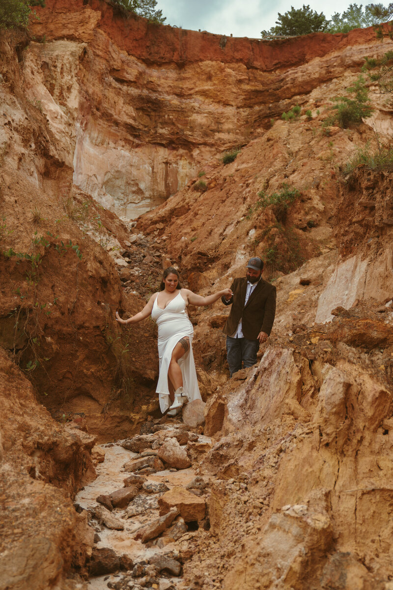Bride in a white dress holding her groom’s hand as they navigate a rugged, earthy canyon landscape during their adventurous elopement