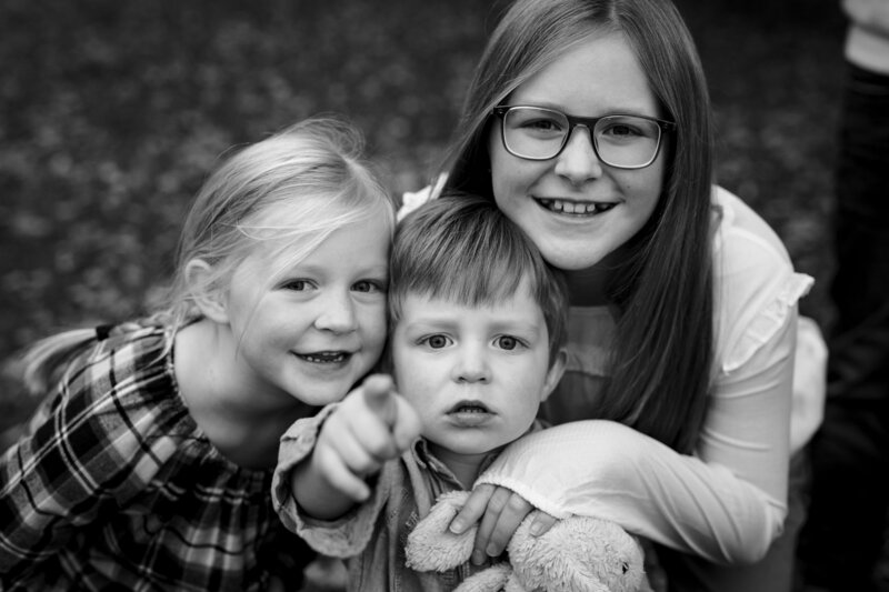 A black and white photo of three children closely grouped together, with a boy in the centre pointing towards the camera. The girls on either side are smiling. They are outdoors at a local Kington park during a family lifestyle photo session.