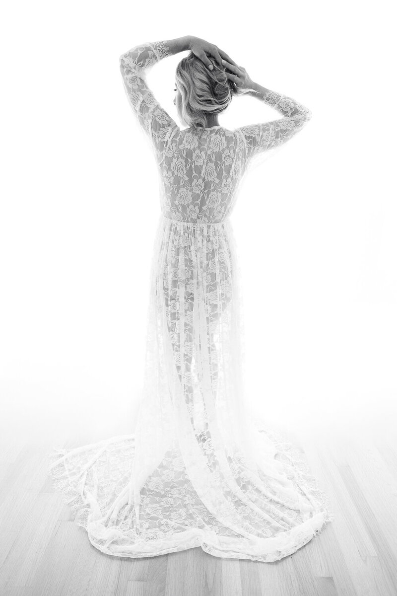 Black and white image of a woman in an Oakland boudoir studio, her figure draped in a flowing white lace gown with a train, standing with her back to the camera. Her hands are raised, elegantly adjusting her hair, as the soft natural light enhances the intricate details of the lace.