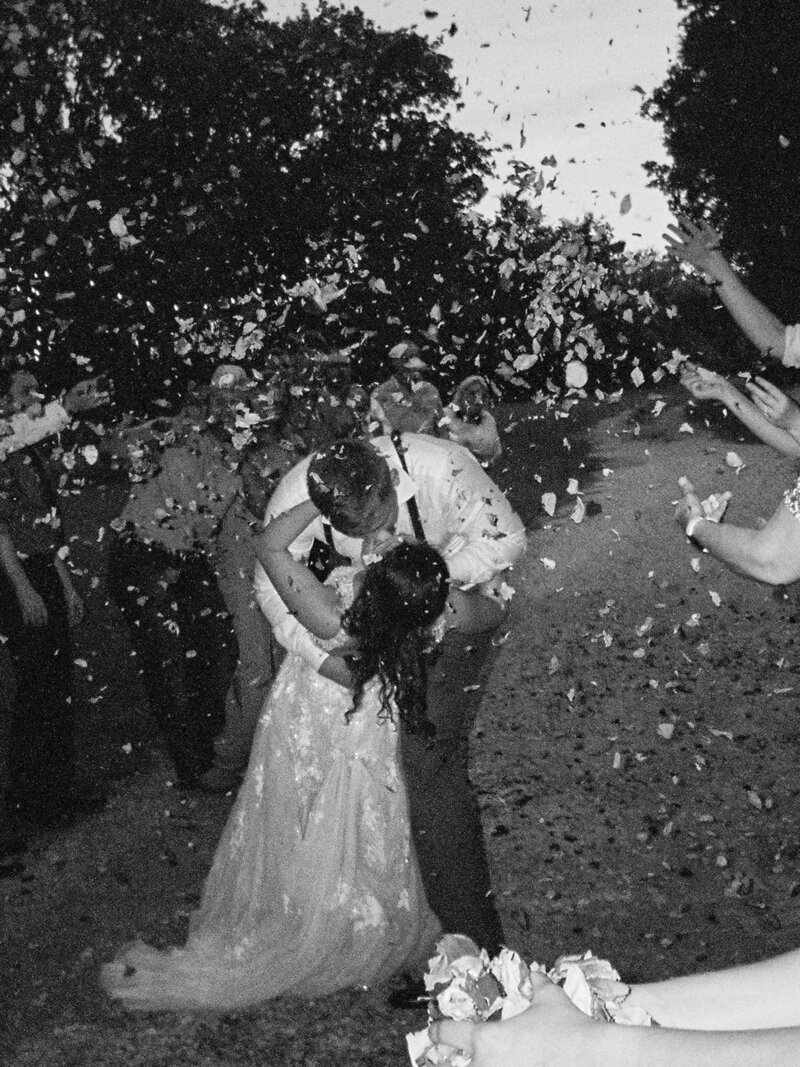 A black and white film photograph of a couple kissing as they leave their wedding reception, with flower petals falling around them.