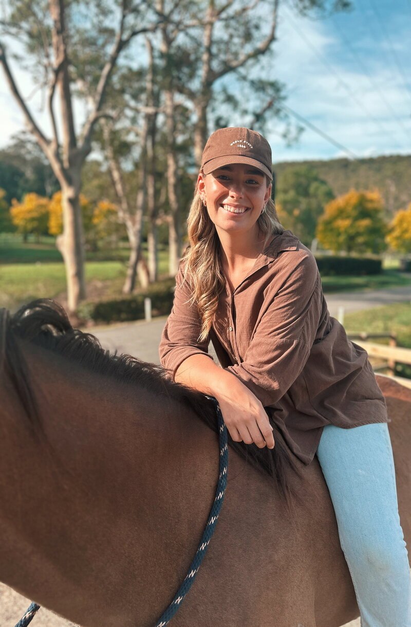 A young boy in a blue top, black pants and a cargo print hat brushing a dark brown horse
