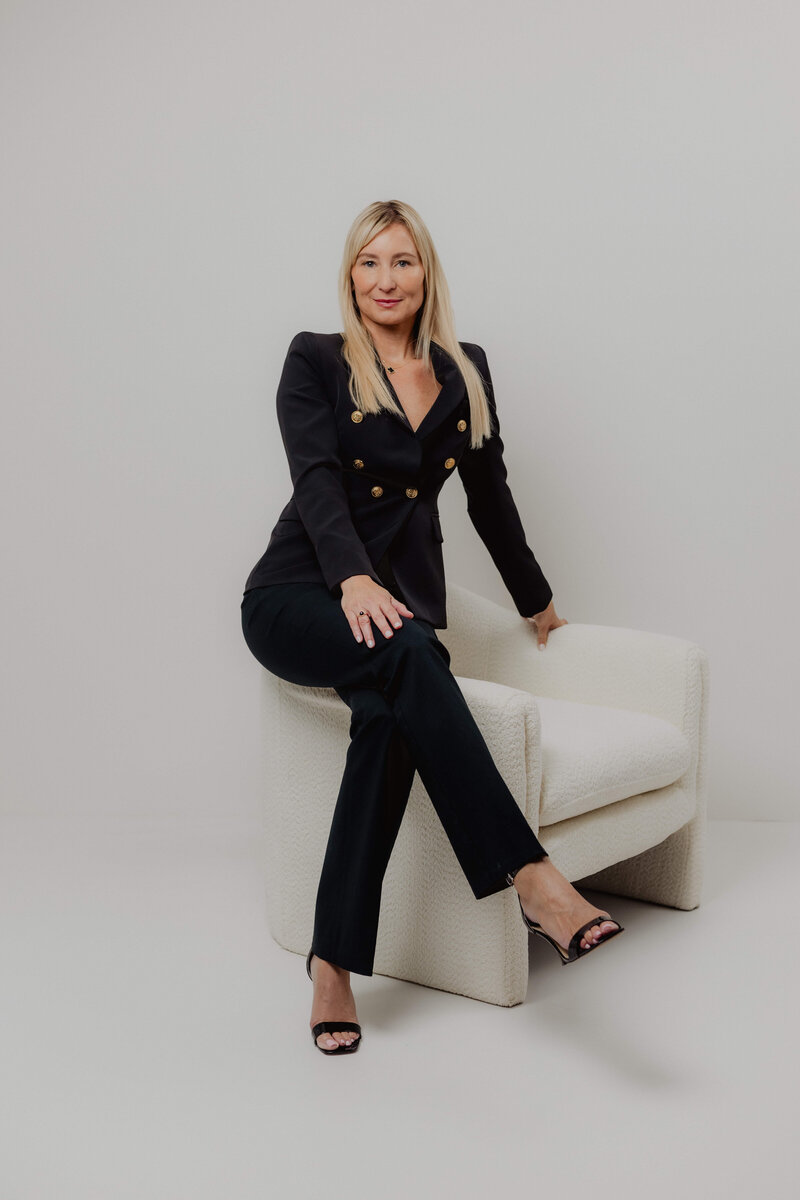 A women sitting on the edge of a white chair with a black suite with her legs crossed. 
