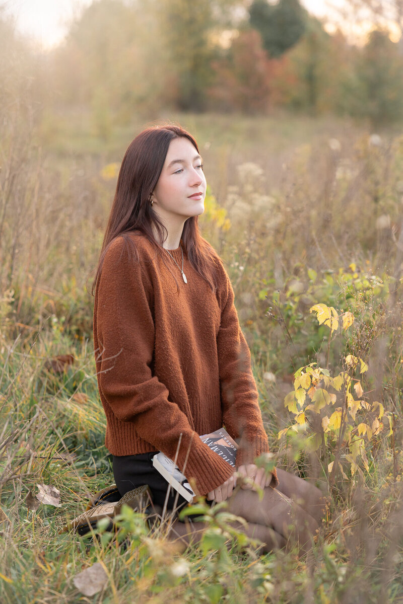 senior girl in an orange sweater and black skirt sits in a field at sunrise with a book in her lap while looking forward to her future, Indianapolis senior photographer
