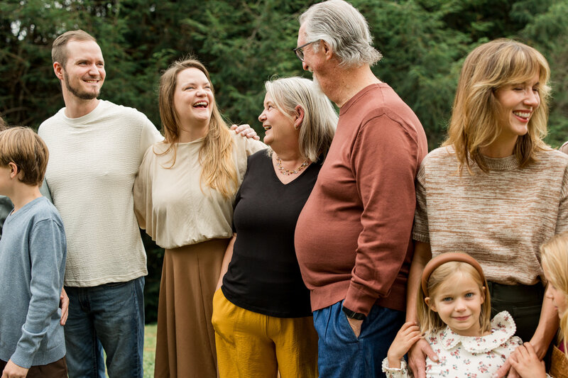 A large family of fifteen leans in and smiles together in front of a stone fireplace all wearing blue, white and cream color coordinated outfits. The grandmother and grandfather are surrounded by their children and grandchildren.