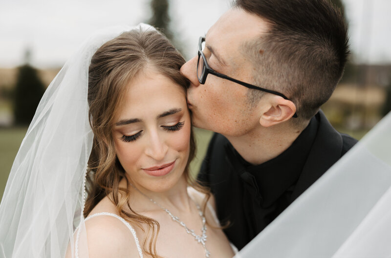 Groom kisses bride on temple and bride looks down soft smile