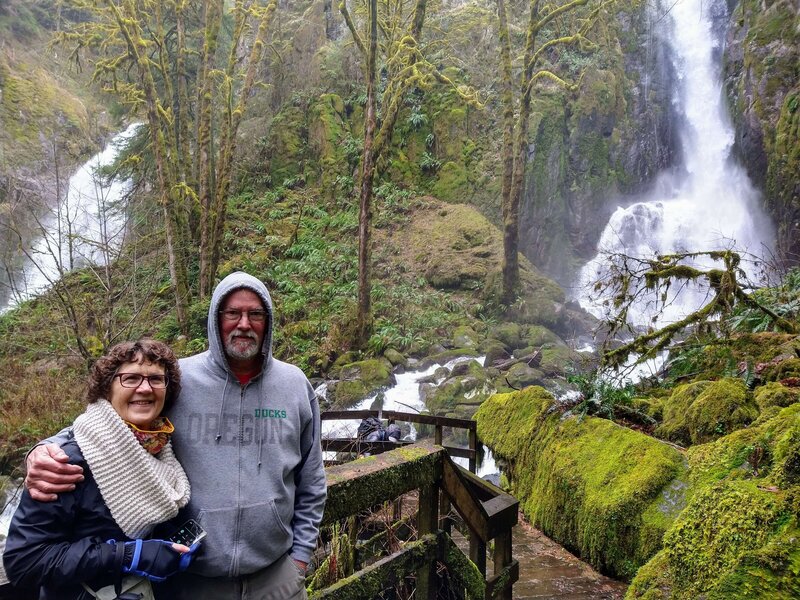 A couple  standing to side of waterfall and lush greenery surrounds them in Oregon.