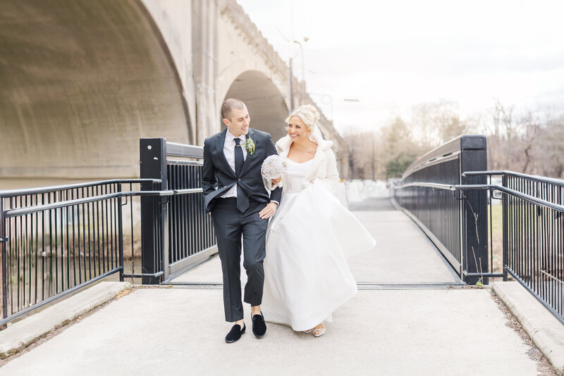 Newlyweds laugh and walk arm in arm under a bridge holding a white bouquet