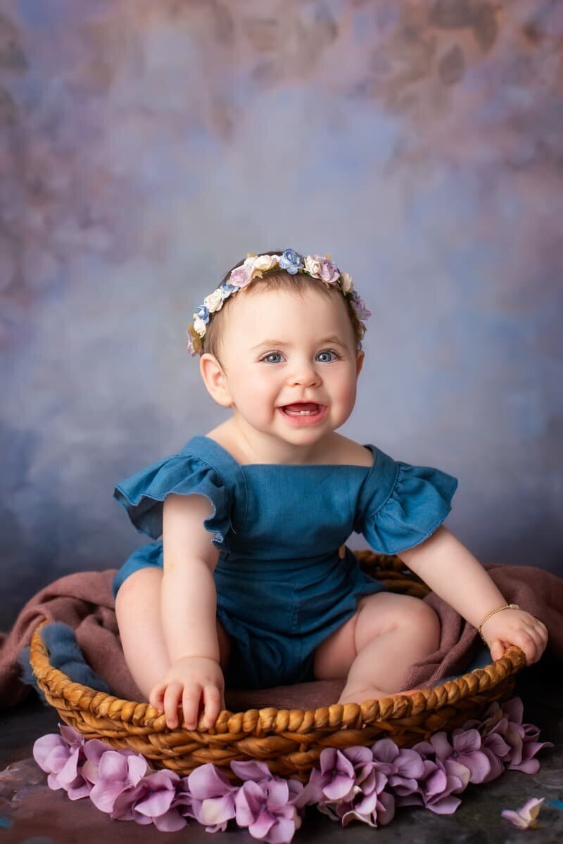Smiling baby girl sitting in a basket wearing a blue romper during milestone session — Birmingham, AL baby photographer.