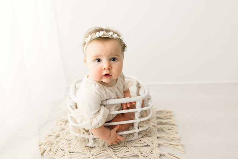 An adorable baby with wide eyes and a cute headband sitting in a small white basket on a cream-colored rug against a bright background.
