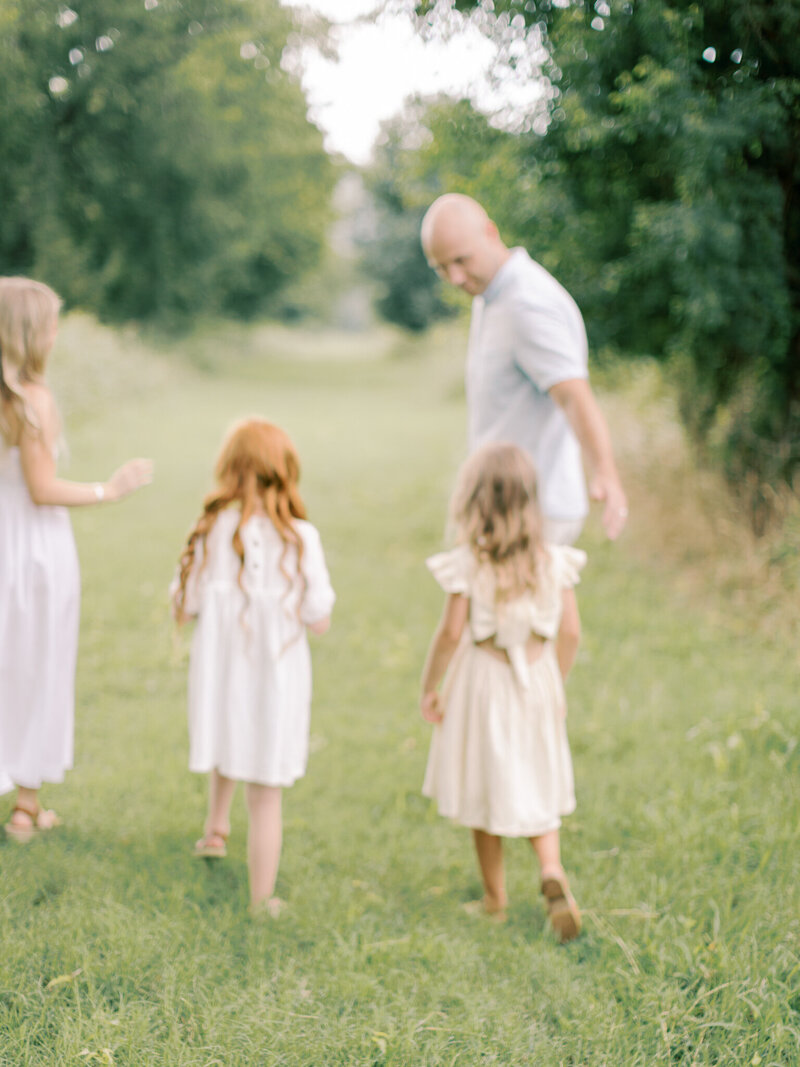 Blonde mother in white dress, red headed daughter with curls in white dress, blonde wavy haired girl in cream dress, and dad in light blue shirt walk away down a grassy path  together taken by Little Rock family photographer Bailey Feeler Photography