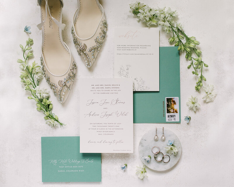 Bridal flat-lay showing beaded high-heels. white flowers, and light teal and white stationery set.