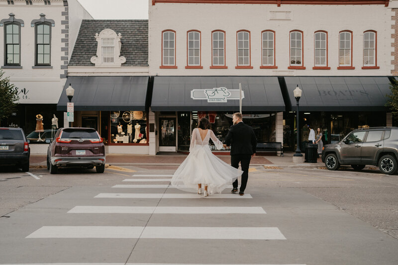 Bride and groom grasp hands as they walk away from the wide angle camera and walk across the street of a small town in Iowa. Brides beautiful white dress blows in the wind as they cross the street happy on their wedding day.