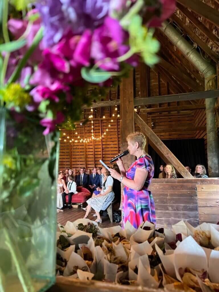 A woman in a colorful dress speaks into a microphone while reading from a paper at a wedding ceremony inside a rustic barn. Guests, including a bride and groom, sit attentively in the background under string lights.