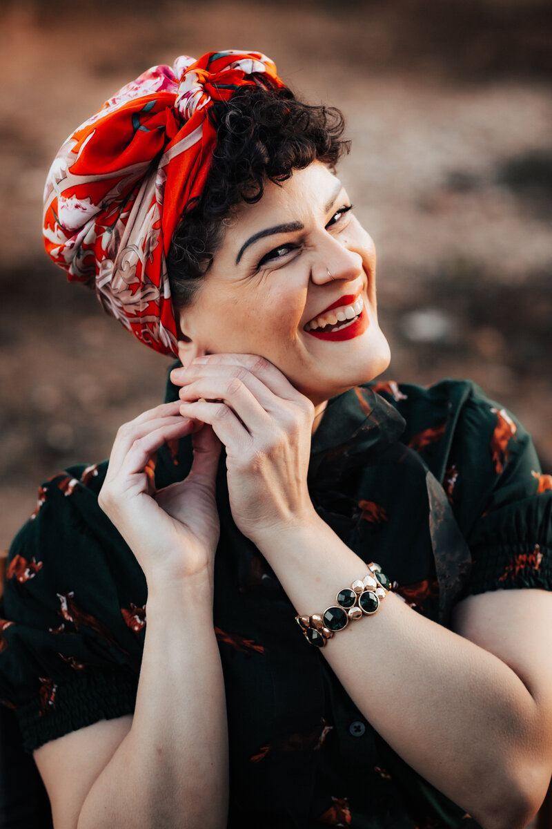 Laura is centered in focus, wearing a dark green shirt dotted with orange foxes, and a red, patterned hair wrap. Her dark, curly hair is visible in the front of the wrap. While adjusting an earring with both hands, Laura smiles brightly at thei viewer.