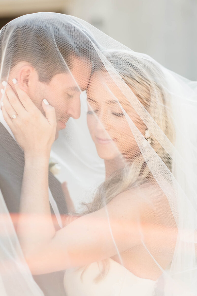A bride and groom share an intimate moment beneath the bride’s delicate veil, softly embracing with eyes closed. The golden light enhances the romantic, ethereal feel of this Alabama wedding photography capture, preserving the love and emotion of their special day.