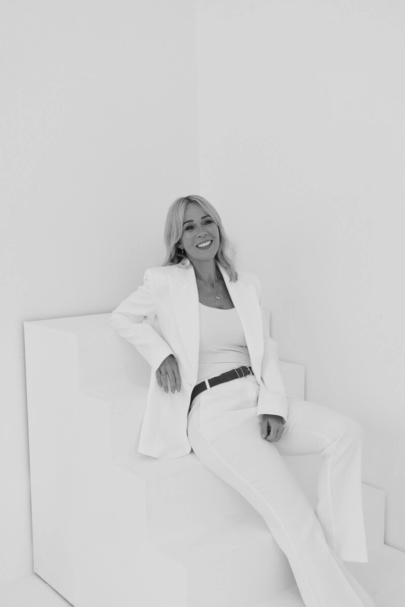 A woman with fair hair, wearing a white suit and vest top, sits smiling on white geometric steps in a bright, minimalist setting.