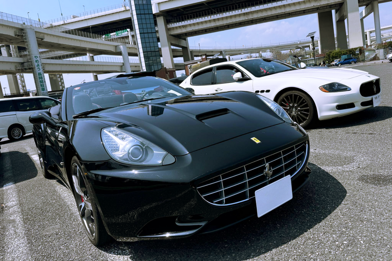 A black Ferrari California and a white Maserati Quattroporte parked side by side under the Tokyo expressway at Daikoku Parking Area, ready for a luxury drive experience.
