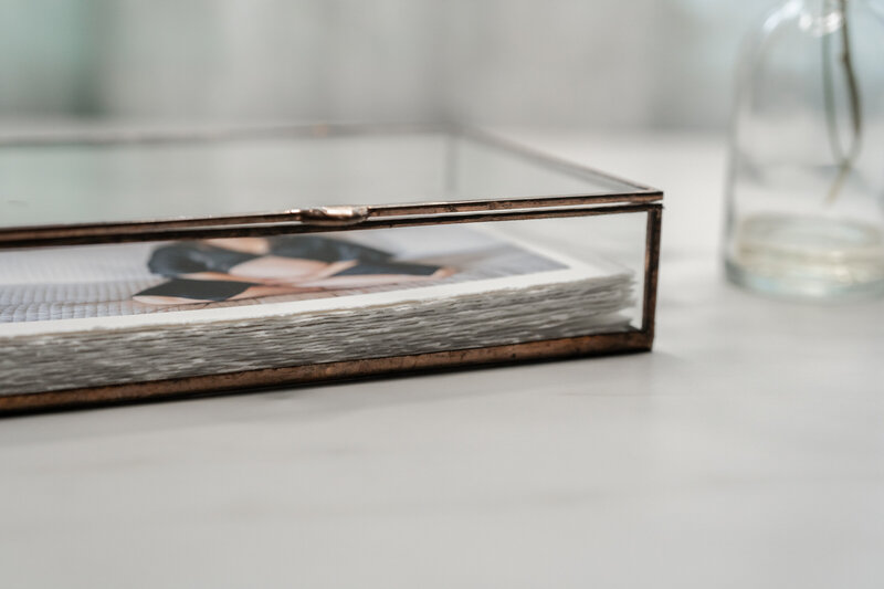 A close-up view of a glass box containing a stack of photo prints, with the top print partially visible, showing a woman in a black outfit.