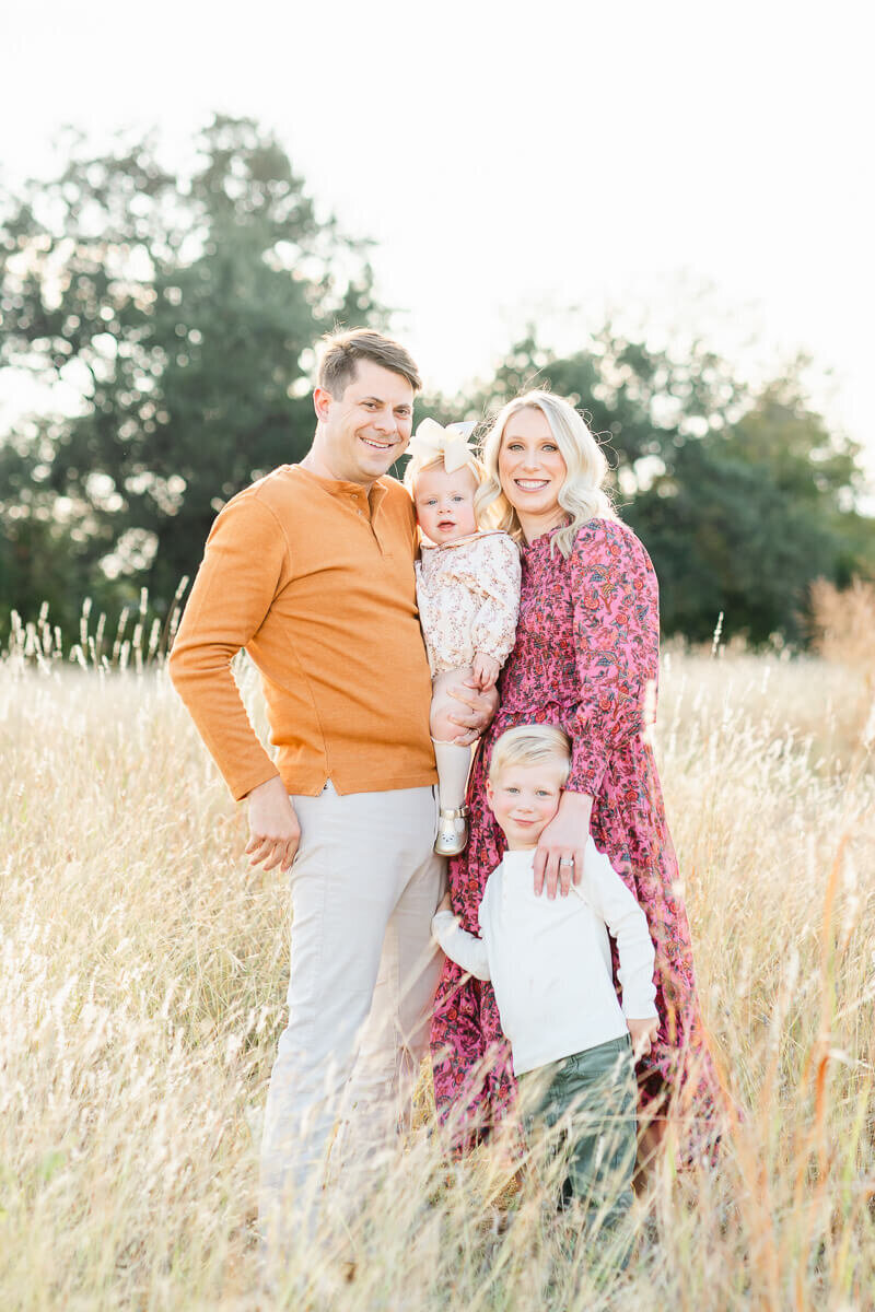 a family of 4 with a mother, father, toddler girl, and little boy stand in a field of tall grass for their fall family pictures in Austin. 