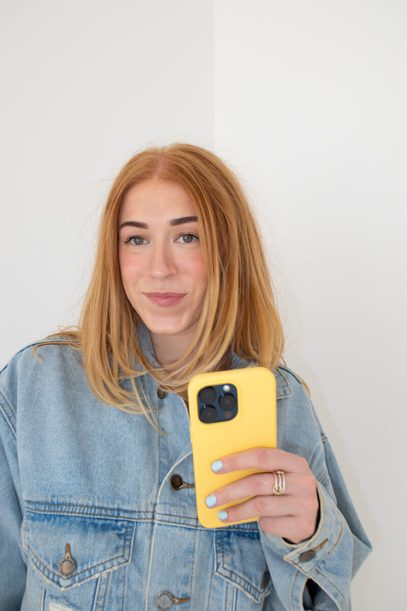 ALT: Grace sitting on floor with arms crossed over knee holding phone smiling at camera with papers on floor under her and taped to wall behind her