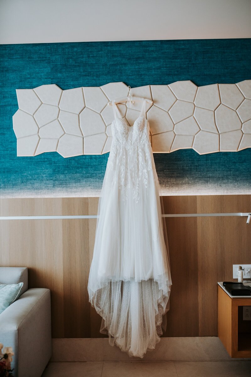 A white lace wedding gown hangs on a wooden wall, beneath a modern geometric art piece, inside a brightly lit room. The gown features a V-neck and a flowing train, displayed against a teal and neutral-toned backdrop.