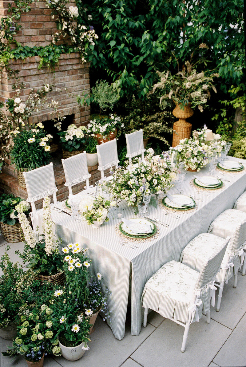 Bride and groom share a kiss on a city street, with the bride in a long white gown and a fur coat, holding a large green and white floral bouquet. Festive garlands and lights adorn the building facade behind them, adding a romantic winter holiday atmosphere to the scene.