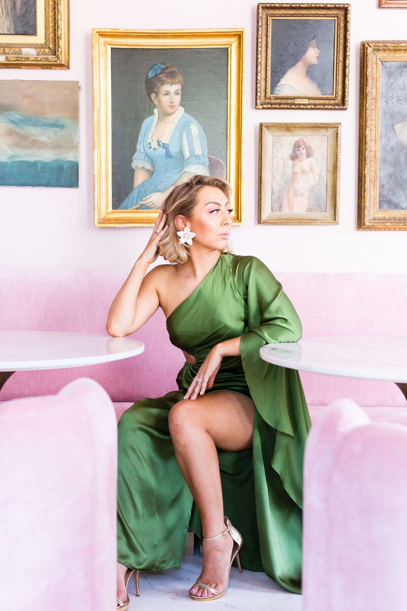 woman wearing green gown sitting on a pink couch and looking away from camera