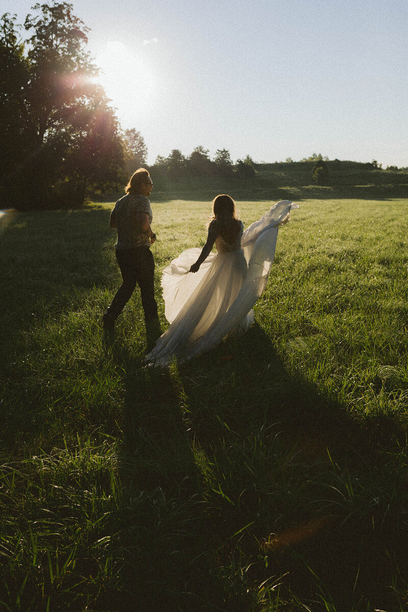 Bride swinging her dress as groom jogs beside her in a large field at sunrise, captured in a dreamy cinematic-style portrait by Emily Ambie.