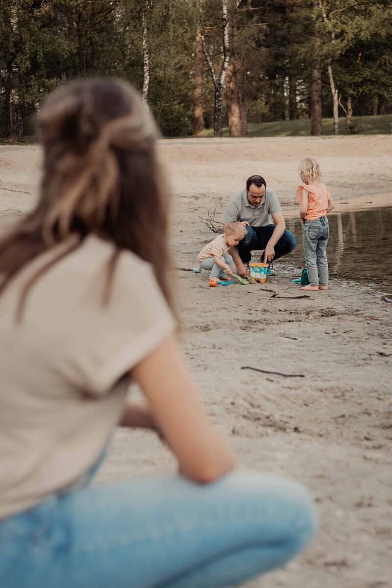 mama kijkend naar papa en haar 2 kinderen die spelen in het zand tijdens de gezinsshoot in zswartsluis