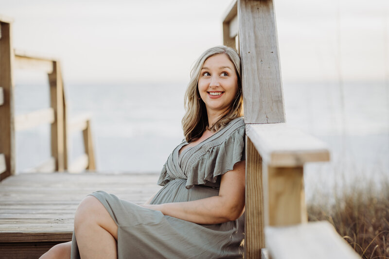 Blonde photographer sits on dock at the beach and smiles during a branding session